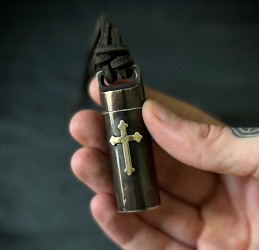 A hand holding a small cylindrical metal pendant or case with a gold cross emblem and a dark cord wrapped around the top, against a dark blurred background.
